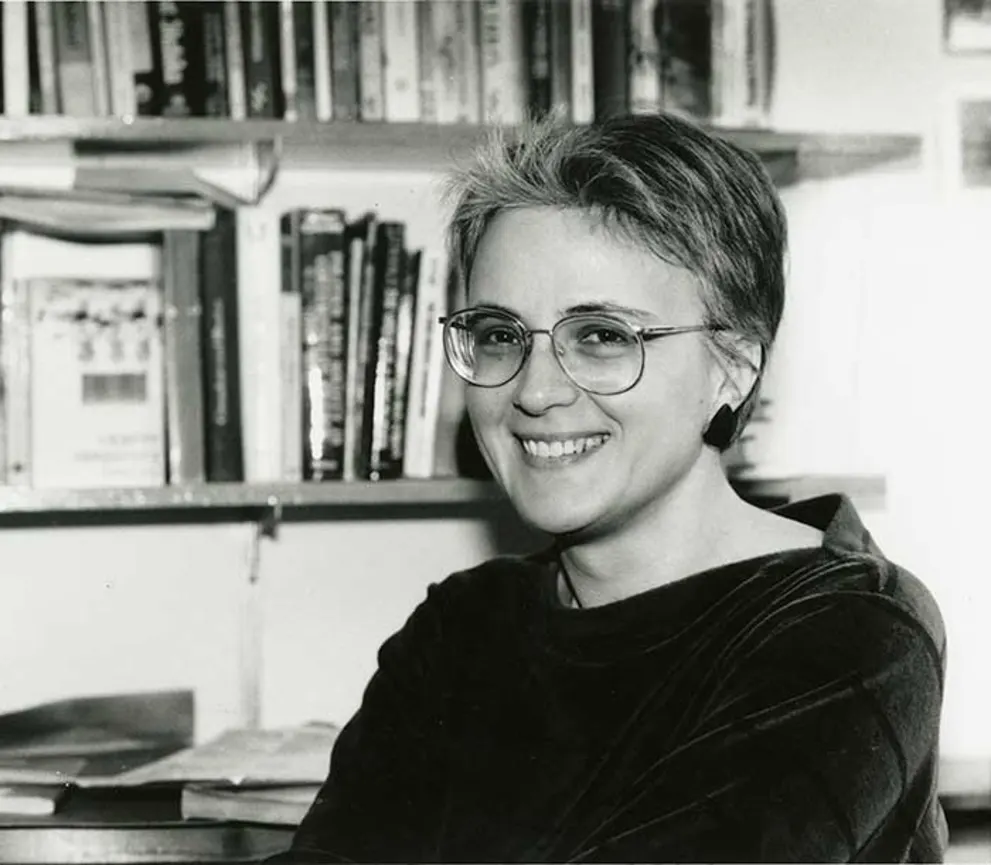 Person smiling, wearing glasses and sitting in front of a bookshelf filled with books.