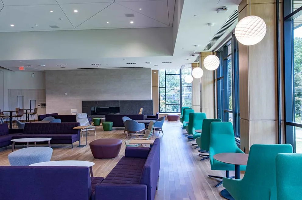 Modern lounge with purple and teal seating, large windows, hanging round lights, and wooden flooring. Tables are scattered throughout the space.