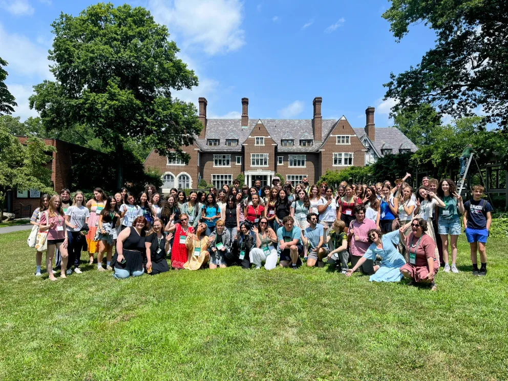 A large group of people posing on a lawn in front of a historic brick building under a clear blue sky with trees surrounding.