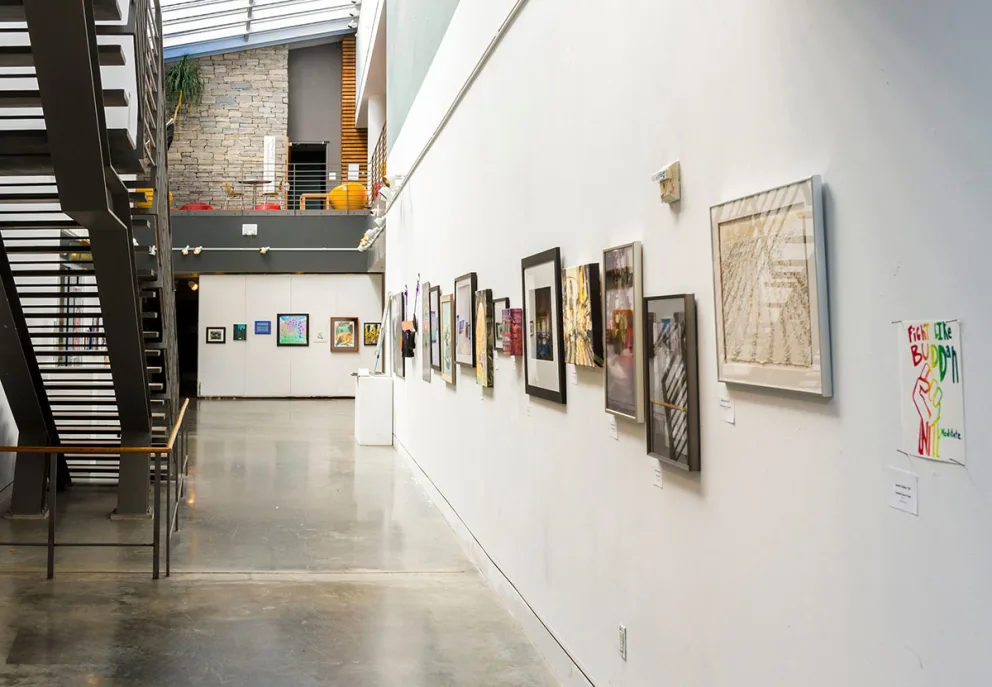 Art gallery interior with framed artworks on a white wall. An industrial-style staircase is on the left. The space is well-lit with natural light.
