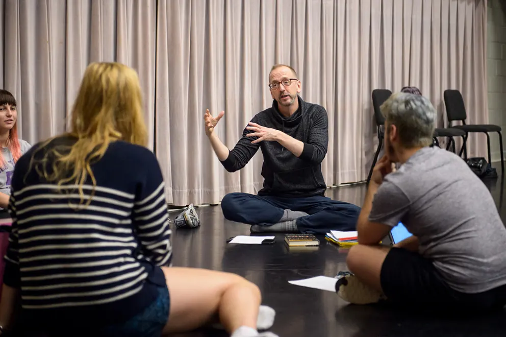 Person sitting cross-legged on the floor, gesturing with hands, engaging in conversation with three seated people in a room with chairs and curtains.