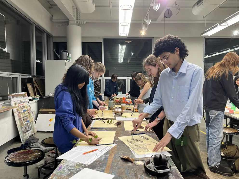 People standing around a table in an art studio, drawing on paper with tools and leaves.
