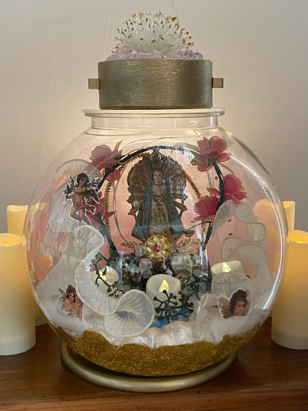 Decorative glass dome with religious figure, flowers, and angel figurines. Surrounded by candles, with a textured object on top.