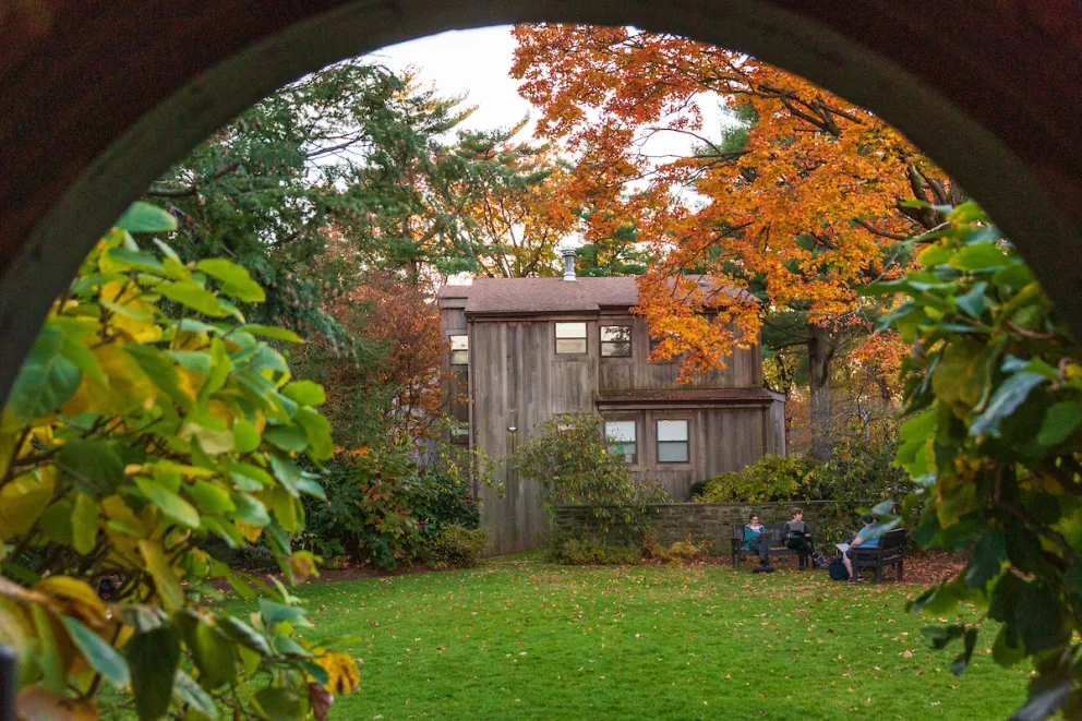 Wooden house framed by autumn trees and foliage, with people sitting on benches in a grassy yard viewed through an archway.