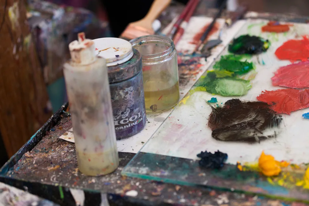 A cluttered artist's table with jars, brushes, and a palette smeared with various colored paints, including red, green, and yellow.