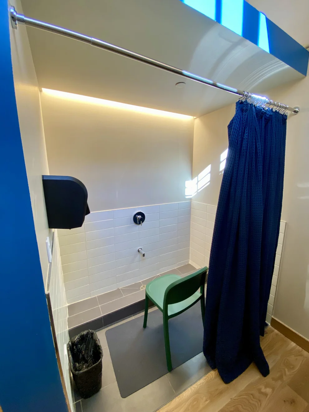 Small shower area with white tiles, a blue curtain, green chair, wall-mounted tap, and garbage bin. Illuminated by overhead lighting.