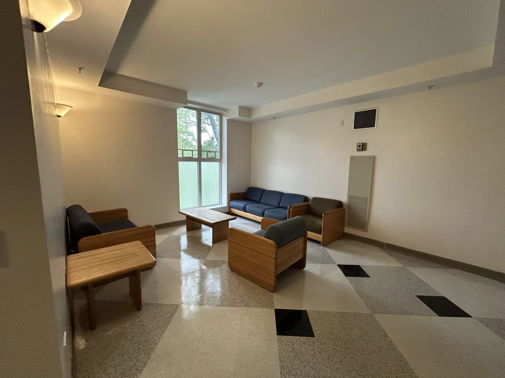 Room with blue cushioned wooden sofas and armchairs, wooden tables, and a large window. Neutral walls and patterned floor.