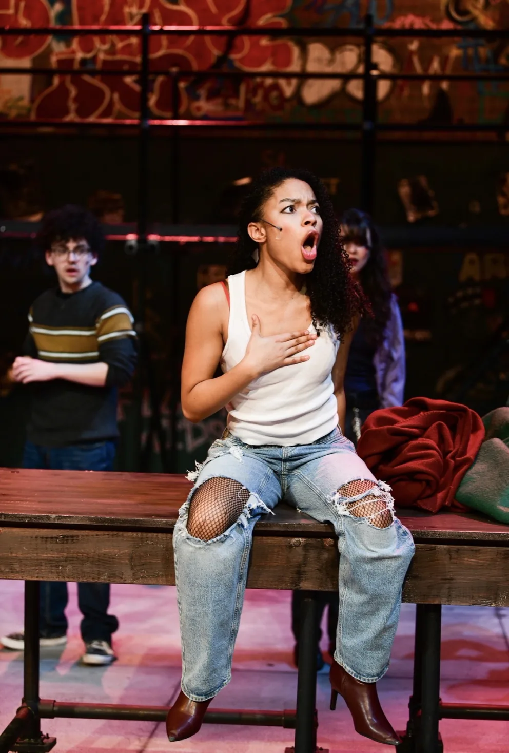 Performer in a white tank top and ripped jeans sits on a table, singing passionately, with two others in the background and graffiti on the wall.