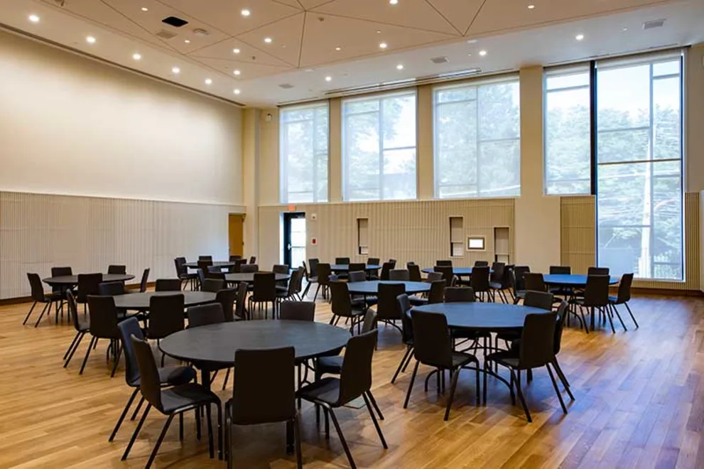 Large modern room with round tables and chairs, wood floor, high ceiling, and tall windows letting in natural light.