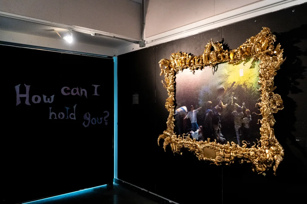 Artwork with elaborate gold frame depicting a lively scene. Text on the wall reads, "How can I hold you?" Subdued lighting.