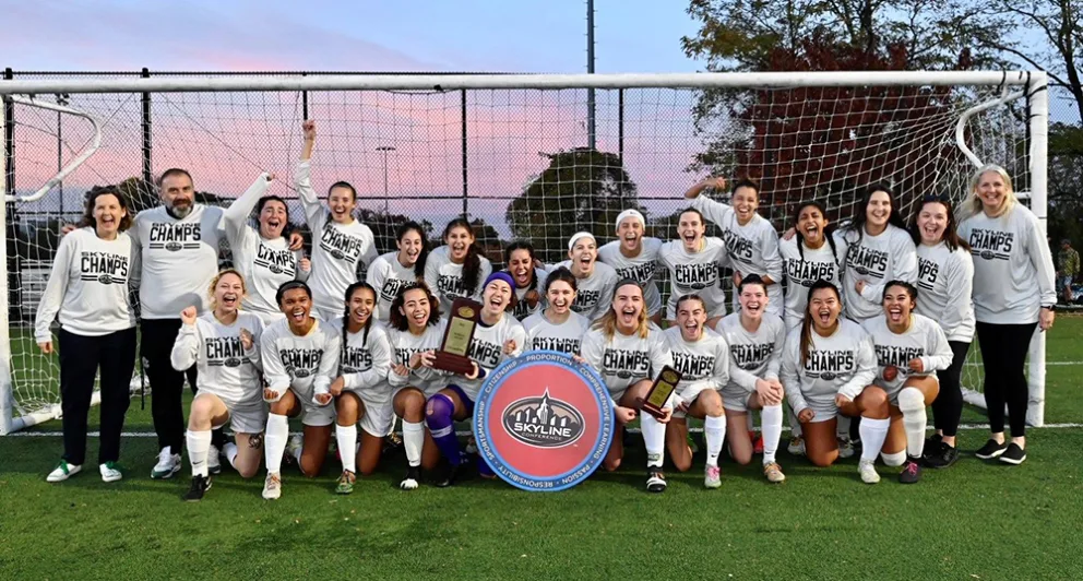 Soccer team celebrating championship victory on field, wearing "CHAMPS" shirts, holding plaques and a "Skyline" sign, standing in front of the goal.