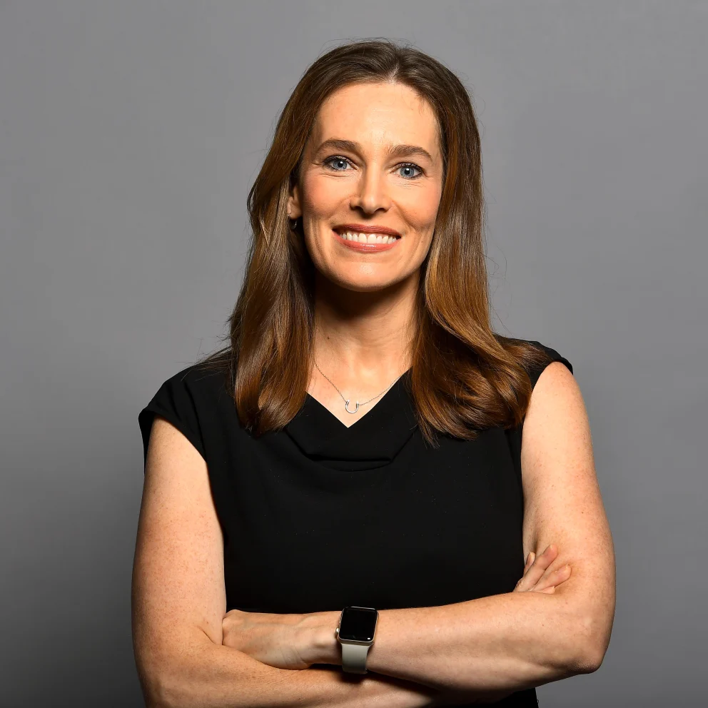 Person with long hair smiling, wearing a black sleeveless top, necklace, and smartwatch, arms crossed, against a gray background.