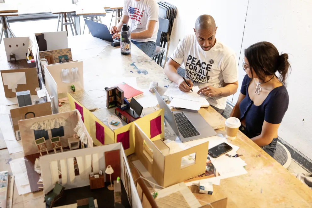 Two people collaborate with models and laptops on a cluttered table, while another person works with a laptop in the background.