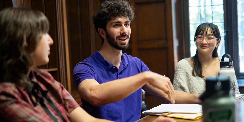 Person in a purple shirt speaks animatedly at a table, engaging with others. Two people listen attentively, with papers and a water bottle nearby.