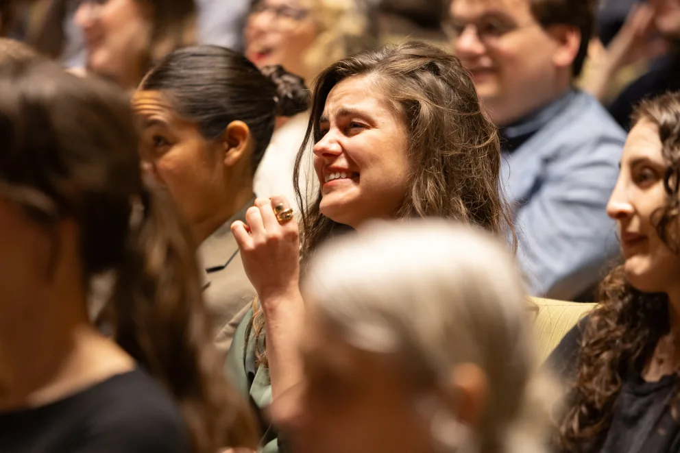 A smiling person with long hair is seated among others in an audience, appearing engaged and delighted.