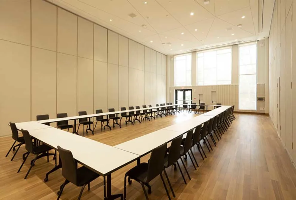 Large conference room with U-shaped table setup, chairs around, wooden floor, and tall windows letting in natural light.