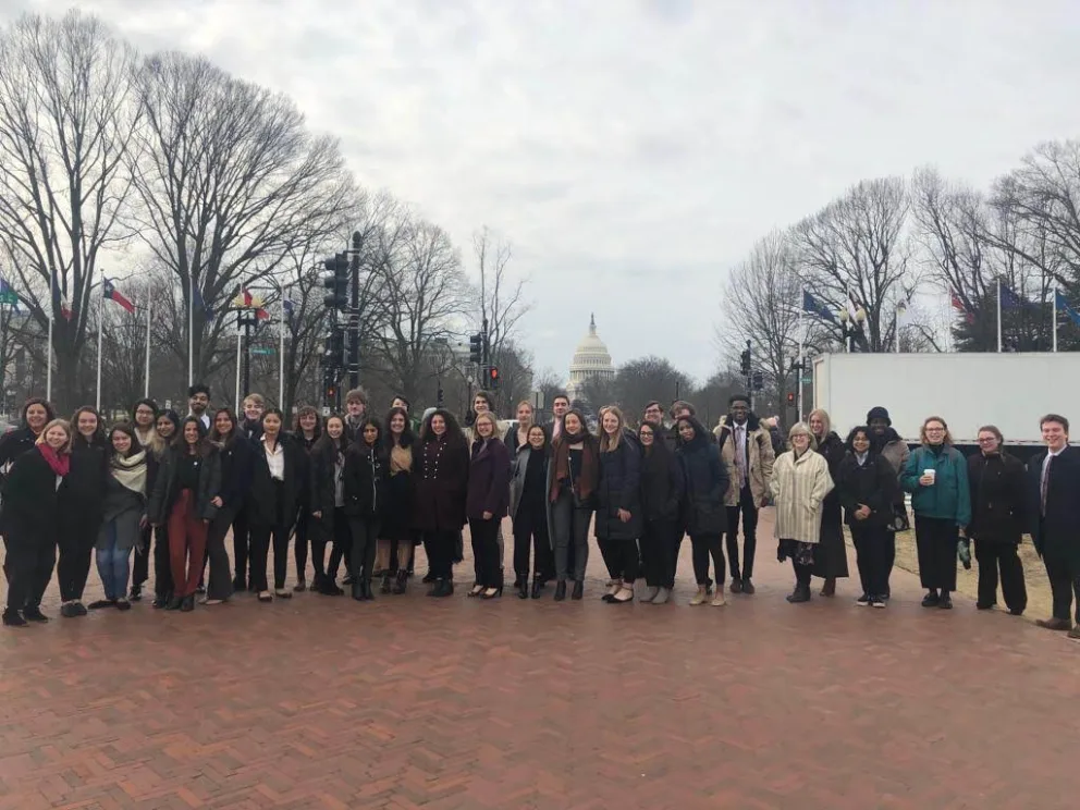 Group of people standing on a brick path with the U.S. Capitol building in the background, surrounded by leafless trees and flagpoles.
