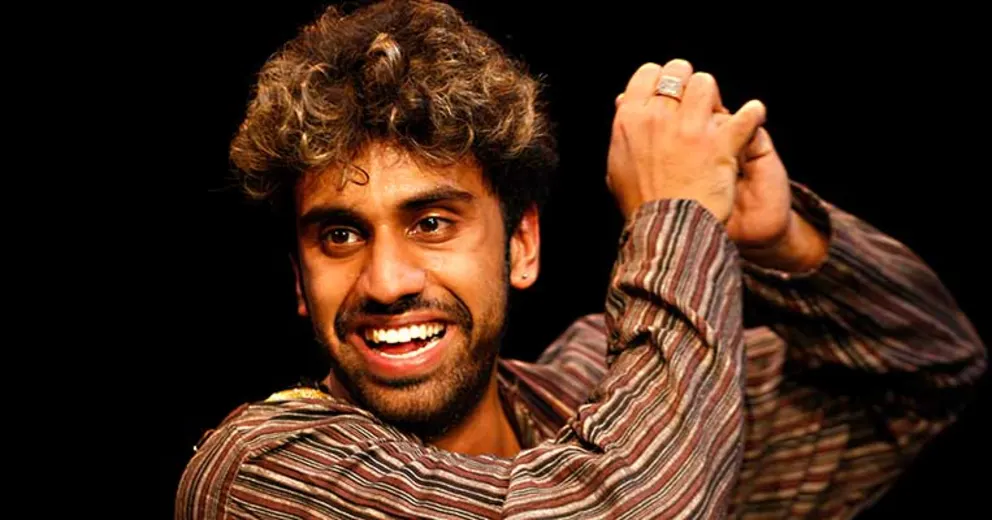 Person smiling broadly, wearing a striped shirt, with curly hair, holding hands up as if clapping or gesturing, against a dark background.
