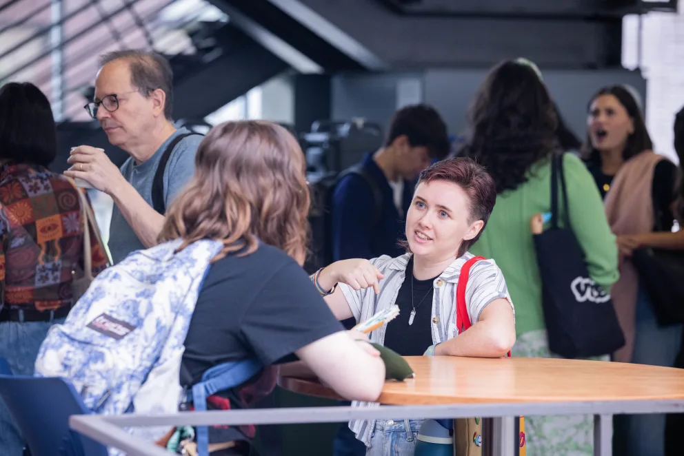 People are gathered in a casual setting. One person is speaking at a round table, while others are engaged in conversation in the background.