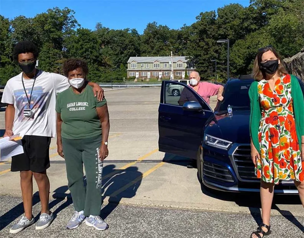 Four people wearing masks stand in a parking lot. Two are near a blue car, one leans on its door, and another wears a floral outfit.