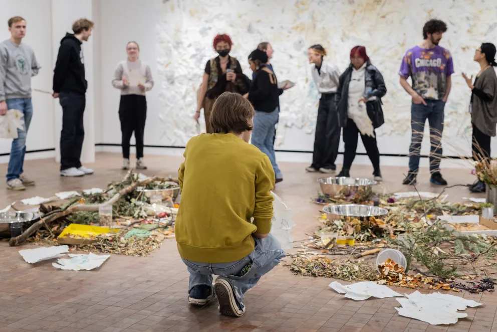 A group of people is gathered in a room, with a man kneeling on the floor surrounded by a pile of papers and other items.