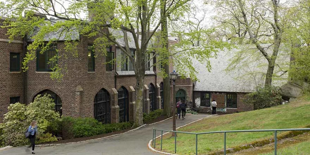 Brick building with large windows, surrounded by trees and grass. A few people walk along the path in front of the building.