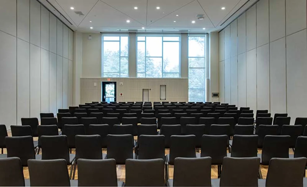 Empty conference room with rows of black chairs facing a large window at the front. High ceiling and lights are on.