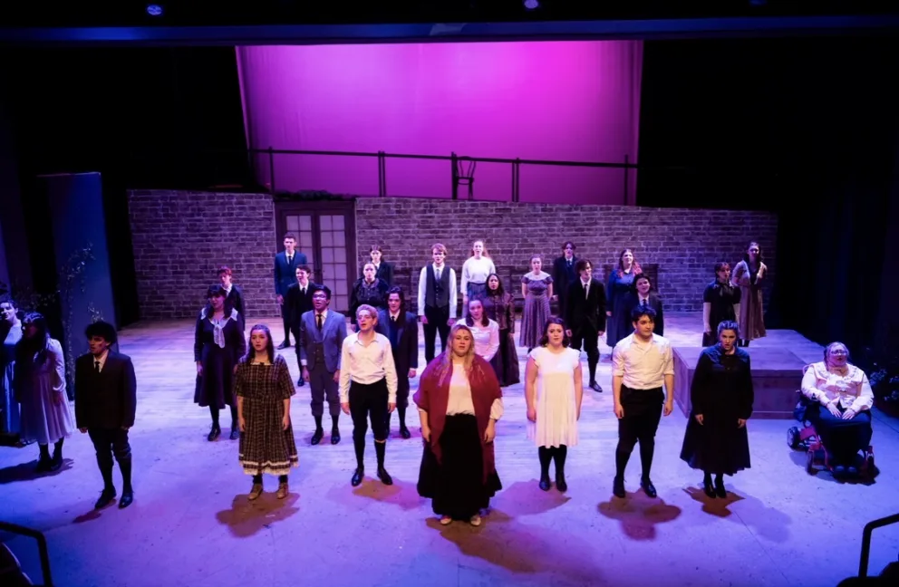A theatrical cast on stage in period costumes stands in formation under purple lighting, with a brick wall backdrop.