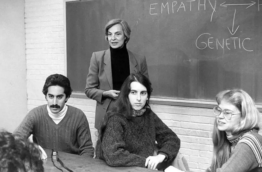 Four people are in a classroom; one stands by a blackboard with "empathy" and "genetic" written, the others sit at a table, facing forward.