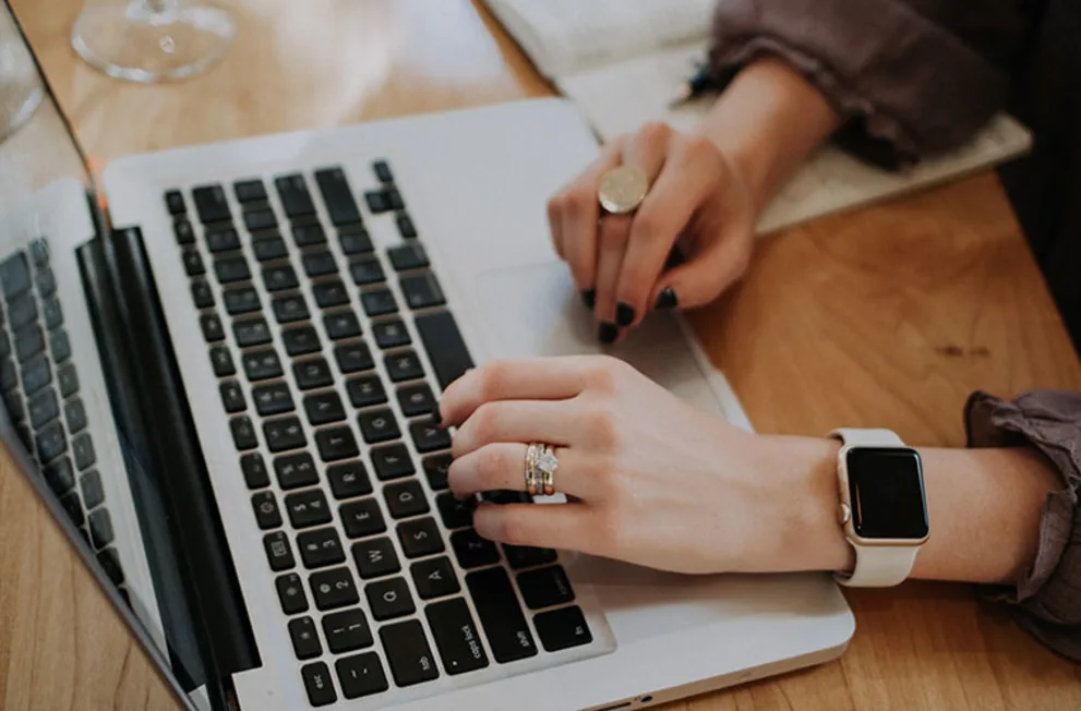Hands typing on a laptop keyboard, wearing a smart watch and rings, with a notebook nearby on a wooden table.