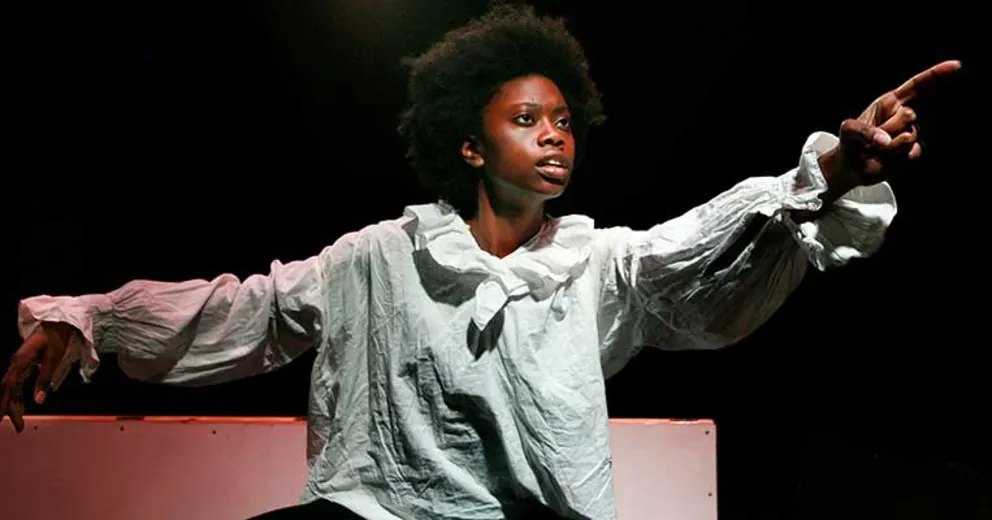 Person in a ruffled shirt gestures dramatically on a dark stage.