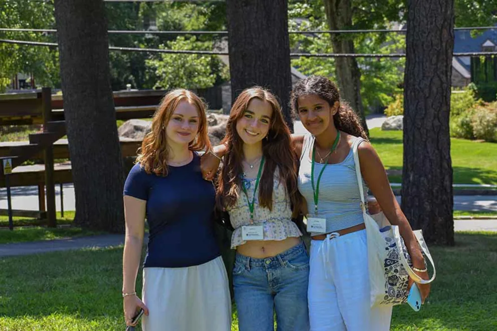 Three people standing on grass, smiling, with trees in the background. Each wears a name tag and has one arm around another's shoulder.