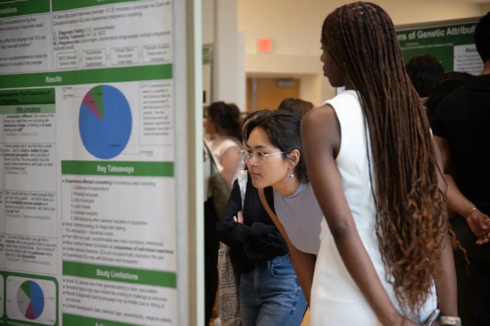 People observing a scientific poster at an event. One individual leans in closely to read the details. The poster includes charts and key takeaways.