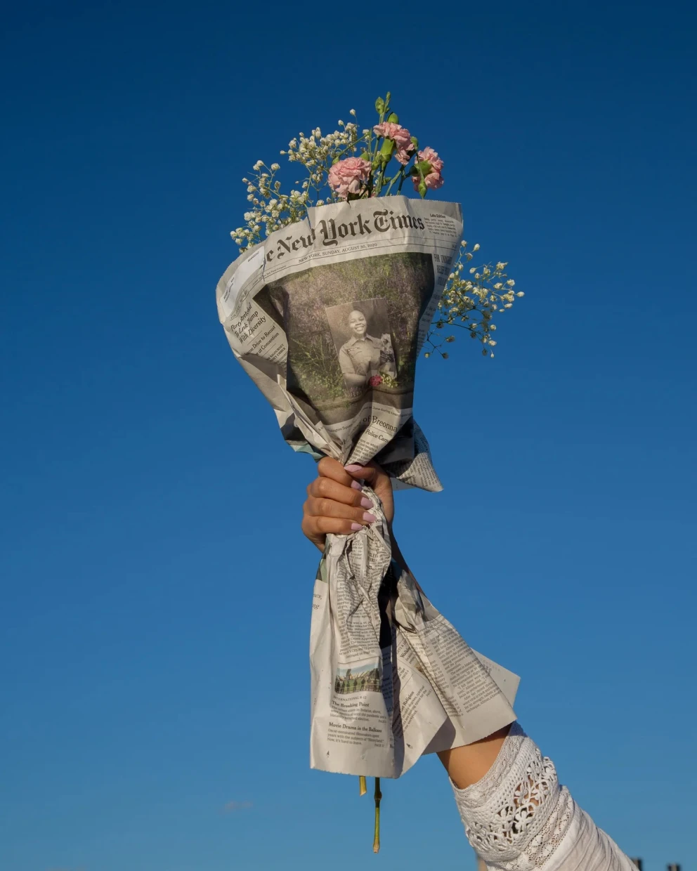 A hand holds a bouquet of pink flowers wrapped in newspaper against a clear blue sky.