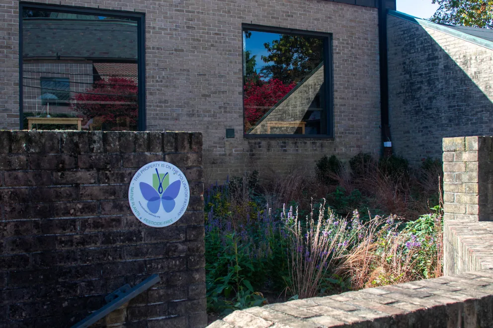 Brick building with a glass window, surrounded by plants and flowers. A circular sign with a butterfly logo is on the brick wall.