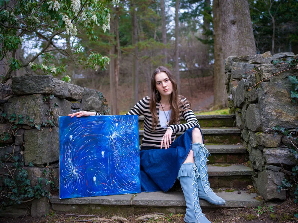 Person sitting on stone steps holding a vibrant blue abstract painting, surrounded by stone walls and greenery in a forested area.