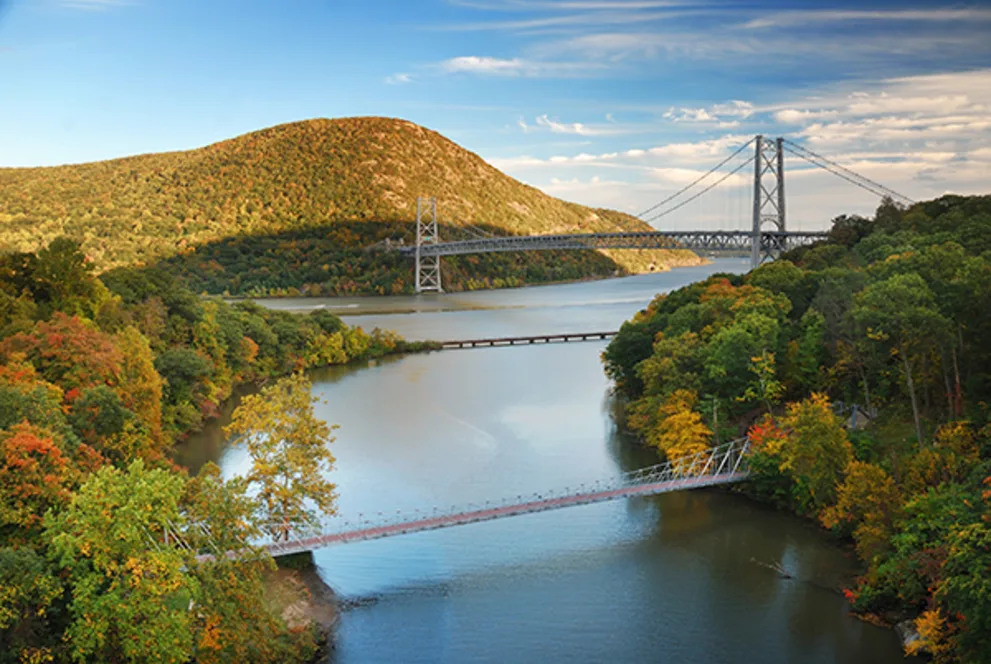 River scene with two suspension bridges, rolling hills, and autumn trees under a clear blue sky with scattered clouds.