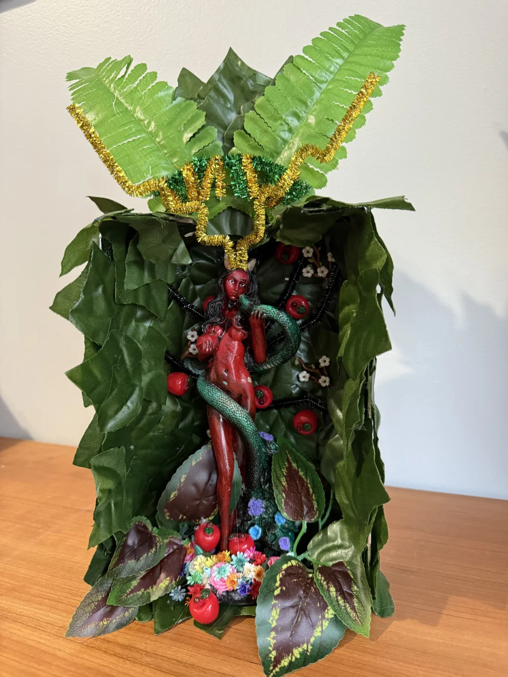 Leafy sculpture with red figure wrapped by a green serpent, surrounded by foliage, red fruits, and colorful flowers on a wooden surface.