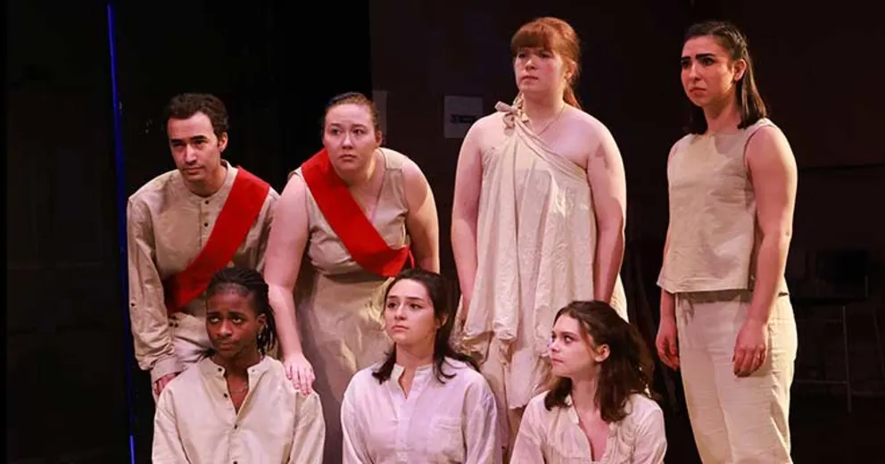 Seven individuals dressed in light-colored, ancient Greek-style clothing pose on stage.