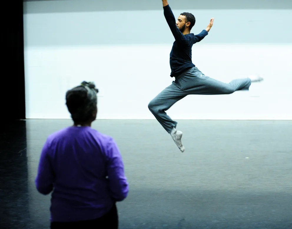A person is captured mid-air in a dance leap on a stage, while another person watches in the foreground, wearing a purple top.