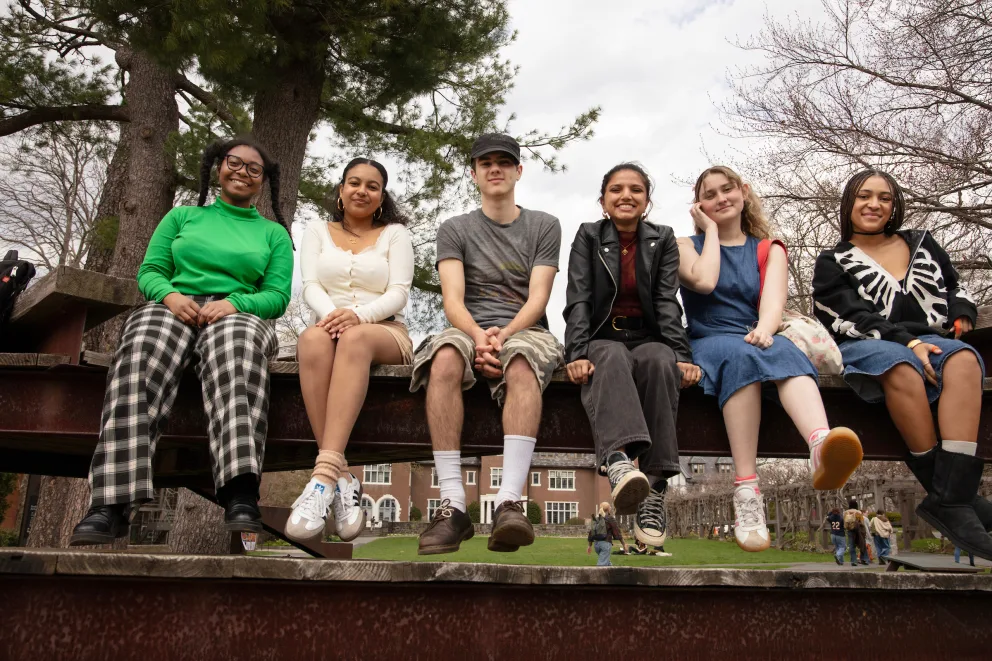 Six people sitting on a railing outdoors, smiling at the camera. Trees and a building are in the background.