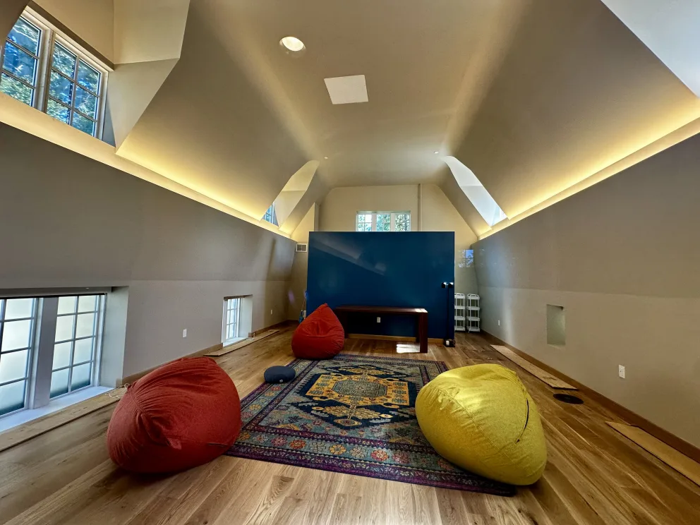 Spacious attic room with wooden floor, colorful bean bags on a patterned rug, angled ceilings, and soft lighting.