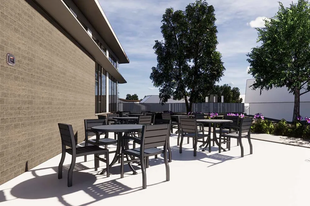 Outdoor patio with several tables and chairs on a concrete surface, adjacent to a brick building. Trees and purple flowers are in the background.