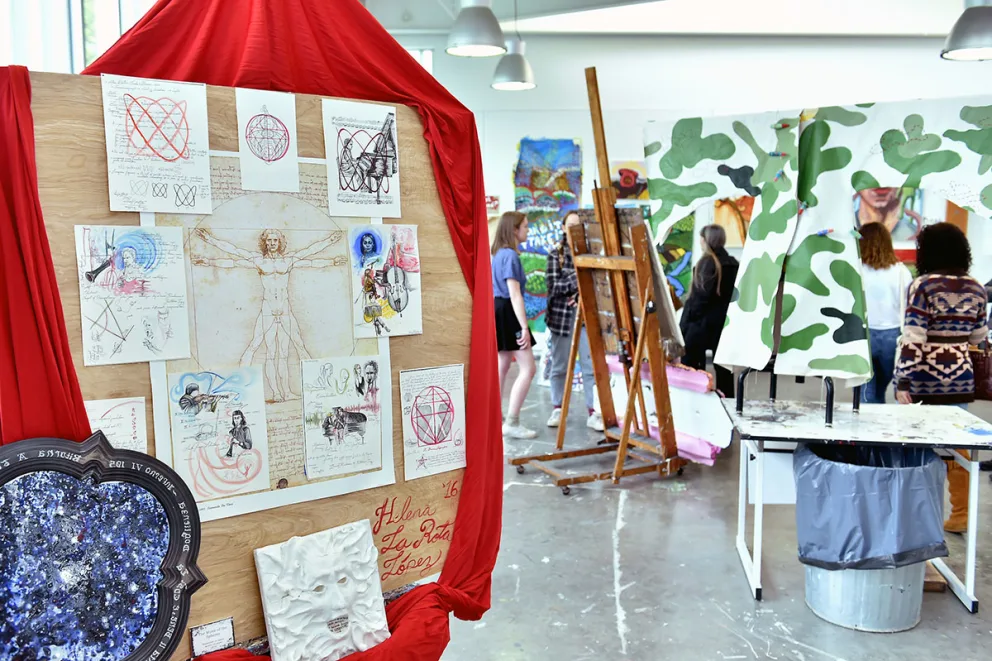 Art studio with paintings and sketches displayed. Easels, artwork, and people are visible, with a large board of drawings in the foreground.