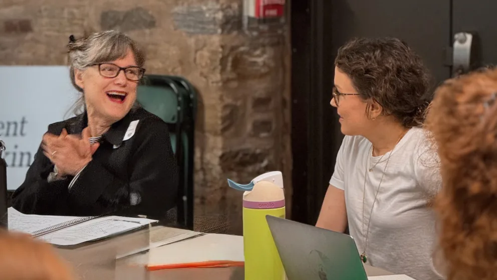 Two people sitting at a table, engaged in conversation. One is clapping, while the other listens. A laptop and water bottle are on the table.