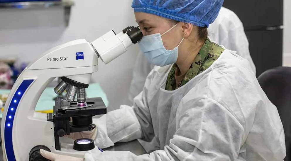 Person in lab coat and mask looks through a microscope in a laboratory.