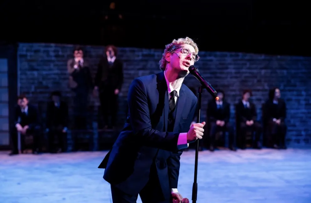 Person in a suit leans forward, singing into a microphone on stage. Brick wall and seated figures in similar attire in the background.