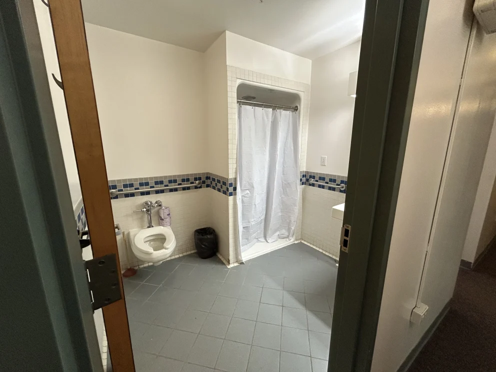 A bathroom with a wall-mounted toilet, a shower with a white curtain, blue-tiled accents, and gray floor tiles. Door is partially open.