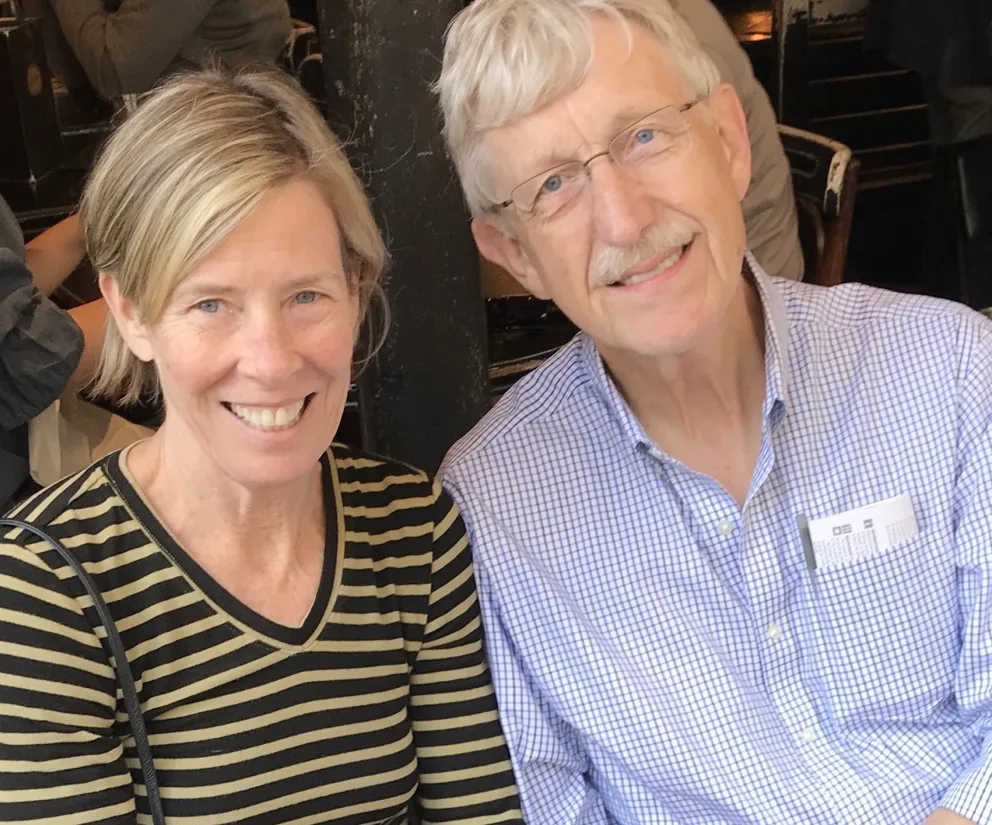 Two people smiling at a cafe table. One is wearing a striped shirt, the other a checkered shirt with glasses, seated close together.