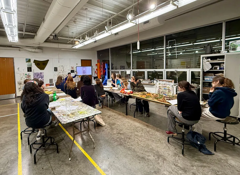 People sit around tables in an art classroom, viewing a presentation on a screen. Walls display artwork, and shelves hold various materials.